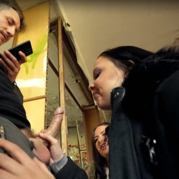 Dark-haired woman in a black jacket giving a blowjob in a hallway while a man holds a phone